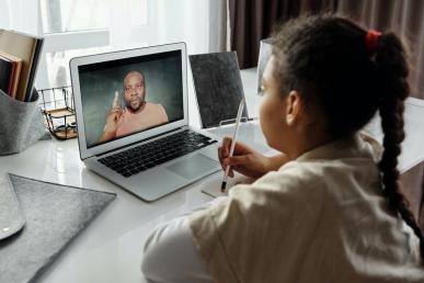 Foto de uma menina negra e de cabelos trançados sentada de costas. Ela segura um lápis enquanto apoia-se em uma mesa repleta de materiais de escritório; inclusive um notebook, no qual ela assiste um homem negro de barba que veste uma blusa salmão e está com um dedo levantado.