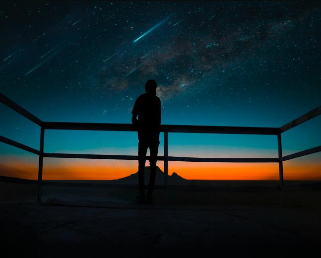 Foto de homem de pé olhando para o céu. Na sua frente, há uma grade que vai até sua cintura, e acima, o céu é estrelado, com algumas estrelas cadentes. O fundo é escuro. 