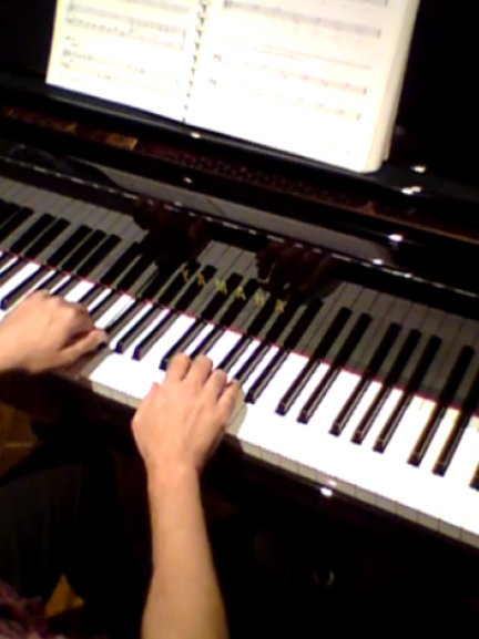 Foto de parte das teclas de um piano sobre o qual estão as mãos de uma pessoa tocando. Na porção superior da imagem está parte de uma partitura apoiada sobre o piano. 