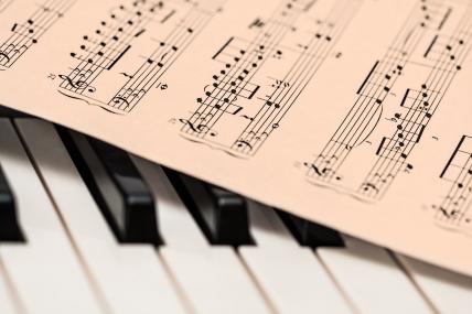 Foto colorida, de detalhe de partitura em cima de teclas de piano.