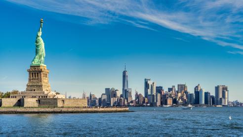 Foto colorida em ambiente externo, da estátua da liberdade e, ao fundo, a cidade de Nova York