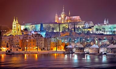 Foto colorida em ambiente externo, de uma noite, mostrando arquitetura de Praga com um castelo ao fundo.