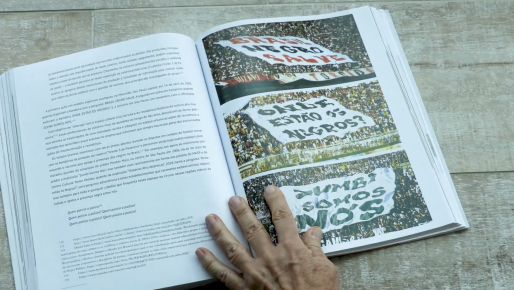 Foto de um livro aberto sobre uma superfície de madeira clara. Na página da esquerda, o fundo é branco e o texto está em letras pretas. Na da direita, há 3 fotos de torcidas organizadas segurando bandeirões com as frases “Brasil negro salve”, “onde estão os negros?” e “Zumbi somos nós”. A mão de uma pessoa branca está apoiada no meio do livro.