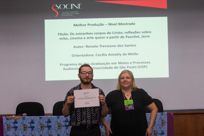 Foto de Renato e sua orientadora Cecília. Renato e Cecília estão de pé, lado a lado. Renato é um homem branco, de cabelos castanhos claros raspados e barba. Ele veste camisa preta com traços brancos e segura o certificado do seu prêmio em mãos. Cecília é uma mulher branca, com cabelos louros médios. Ela veste camiseta preta e tem um crachá verde e roxo pendurado no pescoço. Em segundo plano, está uma mesa com toalha azul e cadeiras pretas. Ao fundo, há uma parede branca com uma projeção com informações sobre a pesquisa de Renato.