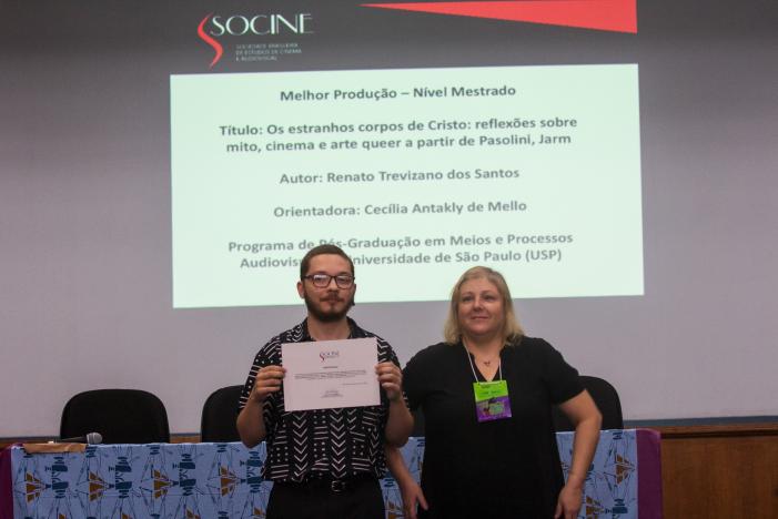 Foto de Renato e sua orientadora Cecília. Renato e Cecília estão de pé, lado a lado. Renato é um homem branco, de cabelos castanhos claros raspados e barba. Ele veste camisa preta com traços brancos e segura o certificado do seu prêmio em mãos. Cecília é uma mulher branca, com cabelos louros médios. Ela veste camiseta preta e tem um crachá verde e roxo pendurado no pescoço. Em segundo plano, está uma mesa com toalha azul e cadeiras pretas. Ao fundo, há uma parede branca com uma projeção com informações sobre a pesquisa de Renato. 