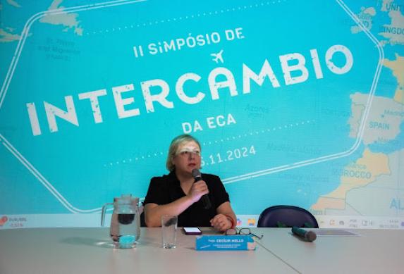 Foto de uma mulher branca sentada atrás de uma mesa branca falando ao microfone. Ela tem cabelos curtos, lisos e loiros e usa uma camiseta preta. Na parede atrás dela, uma projeção em azul com o título “ II Simpósio de Intercâmbio da ECA”, com a ilustração de um mapa. Na mesa, à frente dela, uma jarra e um copo d'água, uma pequena placa azul, óculos, um celular e um microfone.