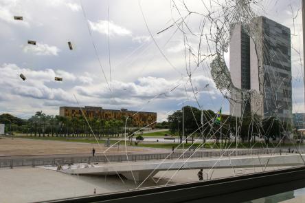 Foto de uma janela quebrada, a imagem foi tirada do lado de dentro do Palácio do Planalto. Através do vidro trincado, é possível ver a rampa do Palácio, uma bandeira do Brasil hasteada, outros prédios, árvores e algumas pessoas.