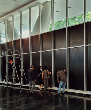 Fotografia colorida mostra o térreo do MAC USP com as janelas pintadas de preto. Enquanto Lucia Koch faz pequenos recortes na superfície pintada, três visitantes observam a paisagem externa através de outros recortes já realizados.