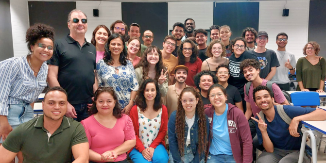 Grupo de cerca de 30 pessoas, em sua maioria brancas e jovens, posam para uma foto. São homens e mulheres, vestindo roupas de diferentes cores. Alguns estão de pé, enquanto outros estão agachados, à frente. Ao fundo, uma parede com listras nas cores branca e preta.