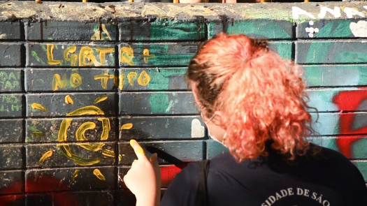 Foto de uma pessoa de costas pintando uma parede. A pessoa é branca, tem cabelos cor-de-rosa cacheados e veste uma camiseta preta. Seus dedos estão sujos de tinta amarela e encostam na parede. Na parede, que é cinza, está a frase “LGBTQ+ no topo” e, embaixo, há o desenho de um Sol, ambos em amarelo.