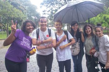 Foto de seis pessoas, dois homens e quatro mulheres, em pé, sorrindo e posando para a câmera. As pessoas estão sujas de tinta roxa e amarela, com as palavras “USP”, “ECA” e “TUR” escritas nos braços e na testa. Uma das mulheres segura um guarda-chuva. Ao fundo, há um gramado e árvores.