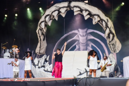 Foto do cantor Jão, suas backing vocals e sua banda durante apresentação no Rock in Rio 2022. Na imagem, o cantor está ao centro usando uma calça cor-de-rosa brilhante, blusa preta e óculos escuros. Ele é branco e tem o cabelo preto. Ao seu lado, duas cantoras de apoio negras usam vestidos brancos e luvas pretas. Elas também usam óculos. Ao fundo, os artistas da banda também aparecem com roupas brancas; um segura um saxofone e outro está tocando bateria. O palco é decorado com elementos que lembram um esqueleto de um animal marinho.