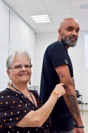 Foto de Silvia Ernestina Soares com Denilson Marques. Ela é uma mulher branca, idosa, de cabelos curtos e grisalhos, tendo uma mecha mais clara na frente. Silvia usa um vestido de mangas curtas preto e com bolinhas, um colar de pérolas, óculos e brincos. Ele é um homem negro, careca, com barba grisalha. Ele veste uma camiseta preta. Silvia aponta para o braço dele, indicando o número 1973 tatuado.