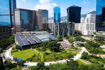 Foto colorida em ambiente externo, de um prédio com placas de energia solar em meio a uma praça.