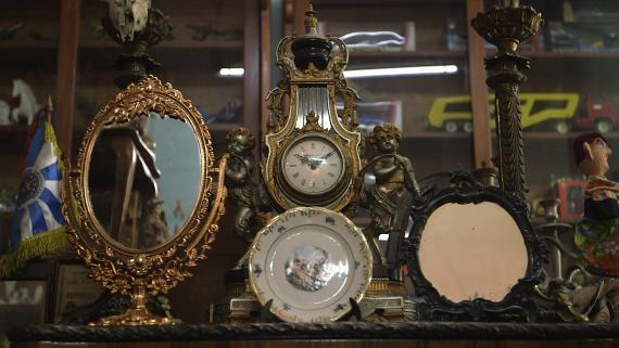 Foto de objetos antigos. Da esquerda para a direita: uma bandeirinha de tecido com listras azuis e brancas e franjas amarelas, um espelho oval com moldura em bronze, um prato com detalhes dourados e pintura no centro, um relógio de ponteiro com dois anjos e detalhes em bronze, um espelho redondo antigo e com manchas e moldura preta, um candelabro antigo e um boneco de tecido vestindo roupas coloridas. Ao fundo, um armário de madeira com brinquedos antigos.