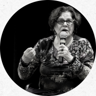 Foto em preto e branco de uma mulher branca, idosa e de cabelos castanhos e curtos. Ela usa óculos, batom e um colar branco. Na mão esquerda, segura um microfone. 
