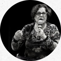 Foto em preto e branco de uma mulher branca, idosa e de cabelos castanhos e curtos. Ela usa óculos, batom e um colar branco. Na mão esquerda, segura um microfone.