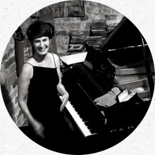 Em uma fotografia preto e branco em formato circular, Ana Fridman, uma mulher branca de cabelos curtos, aparece sorridente ao lado de um piano