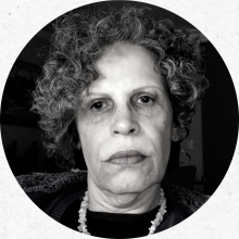 Foto do rosto da professora Irene Machado. A foto está em preto e branco com layout circular. Uma mulher mais velha de expressão séria, cabelos cacheados curtos e grisalhos na altura das orelhas. Ela está usando um colar de várias pedras brancas