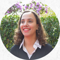 Na foto, uma mulher de cabelos castanhos e ondulados acima dos ombros sorri. Ela veste blusa azul escura sobre camisa com gola branca e listras vermelhas. Ao fundo, arbustos verdes e flores roxas desfocadas.