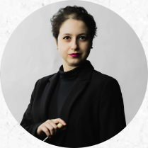 Na foto, uma mulher branca de cabelos castanhos e curtos e olhos escuros segura uma batuta. Ela veste blazer preto, blusa de gola alta também preta e usa batom vermelho. O fundo é branco.