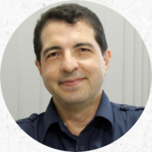 Foto de um homem branco de cabelos pretos, escuros e curtos e olhos castanhos, esboçando um sorriso. Ele veste uma camisa azul marinho. O fundo é branco.
