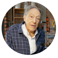 Foto de homem branco idoso. Seus cabelos são grisalhos e cacheados. Ele usa óculos de armação oval fina e uma camisa azul.