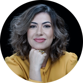 Foto de uma mulher branca sorrindo. Ela tem cabelos ondulados e castanhos com luzes, na altura dos ombros, olhos castanhos, veste uma blusa amarela de manga comprida, usa um anel dourado na mão, apoiada sob o queixo. O fundo é preto.