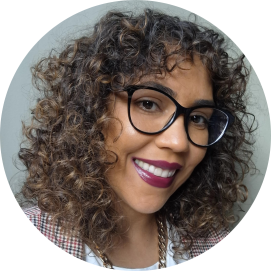 Foto de  uma mulher negra sorrindo. Ela tem cabelos cacheados e castanhos, na altura do ombro, olhos castanhos, veste blusa branca com terno xadrez preto, vermelho e branco, usa uma corrente dourada, batom e óculos de armação preta. O fundo é verde claro.