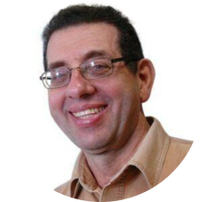 Foto de um homem branco. Ele tem cabelos curtos e escuros, olhos claros, veste uma camisa bege, um óculos de armação preta e sorri. O fundo é branco.