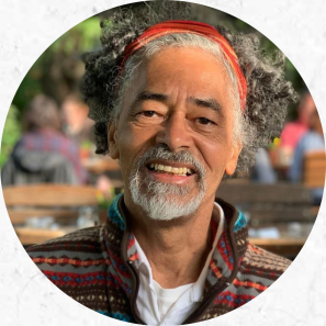 Foto de Almir Almas. Um homem de pele negra, olhos escuros, cavanhaque e cabelos cacheados, curtos, e grisalhos, que estão puxados para trás por uma faixa vermelha. Ele sorri e veste um casaco com listras e detalhes coloridos por cima de uma camisa e uma camiseta brancas. Ao fundo pessoas em desfoque.