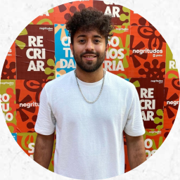 Fotografia de  um homem negro de pele clara, cabelo e barba curtos, enrolados e castanhos. Ele veste uma camiseta branca e usa uma corrente prateada no pescoço. No fundo, cartazes em laranja, verde, marrom e azul com o texto “Recriar”.