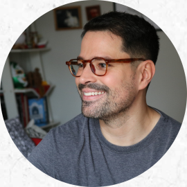 Retrato de um homem sorrindo e levemente direcionado para a diagonal esquerda da imagem. Ele é branco, tem barba e bigode curtos e pretos, cabelos curtos e escuros e usa óculos marrom e camiseta cinza. O recorte da foto vai até a altura do peito e atrás dele há uma parede branca com quadros de tamanhos variados e uma estante com diversos objetos.