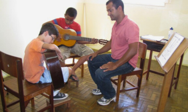 Foto de dois estudantes e um professor sentados em uma sala. Os estudantes,uma criança branca e um adolescente negro usando camiseta e  bermuda, dedilham violões enquanto o professor observa. Ele é negro, tem cabelos curtos e escuros, usa camisa polo e calça jeans. Atrás dele há uma mesa pequena de madeira sobre a qual há folhas de papel e uma garrafa plástica. Ao lado dele há um suporte de madeira para partituras com algumas folhas de papel. O piso é de madeira e ao fundo há uma janela aberta por onde entra muita claridade. 