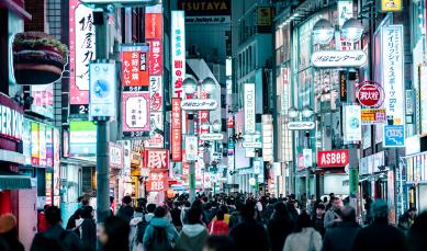 Foto colorida, em ambiente externo, à noite, de rua em Tóquio com vários outdoor coloridos e multidão caminhando
