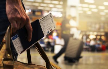 Foto colorida, em ambiente interno de aeroporto, mão segura bagagem e passaporte com bilhete