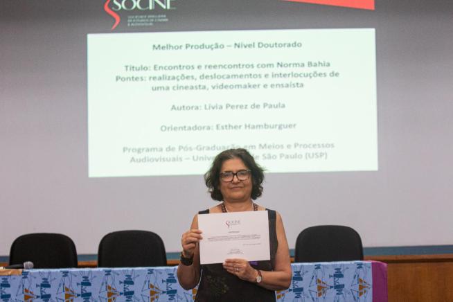 Foto de Esther Hamburguer segurando o certificado de premiação da Socine. Esther é uma mulher branca, de cabelos médios, castanhos e um pouco grisalhos. Ela usa óculos, relógio de pulso, pulseiras, colares e blusa regata preta. Em segundo plano, está uma mesa com toalha azul e cadeiras pretas. Ao fundo, em uma parede branca, estão projetadas informações sobre a premiada, Lívia Perez, e sua pesquisa.