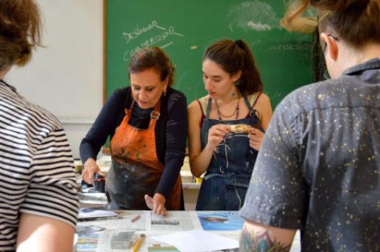 Na fotografia, duas mulheres brancas estão lado a lado. A mulher à esquerda tem cabelos castanhos avermelhados presos, usa avental laranja com respingos de tinta azul e uma blusa azul de mangas compridas. Ela segura um ferro de passar acima de uma mesa forrada com jornais. A mulher à direita tem cabelos lisos e castanhos presos em um rabo de cavalo. Ela usa um avental jeans, uma regata vermelha e colares. Ela segura um objeto pequeno e olha para a mesa. Em primeiro plano, em cada lateral da imagem há uma pessoa de costas.