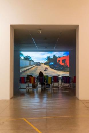 Foto de sala de vídeo exibindo filme. Há um conjunto de cadeiras de praia na sala, e uma mulher está sentada no centro da sala. A projeção mostra uma sala por onde passam carros.
