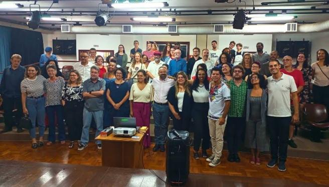 Foto de um grupo heterogêneo de cerca de 50 pessoas em pé em uma sala. Há homens e mulheres de meia idade e alguns jovens, todos usam roupas coloridas, alguns à frente e alguns atrás, em um patamar mais alto. Ao fundo há uma parede branca, o chão é de tacos, no teto há refletores e dutos de ar condicionado e à frente do grupo há uma pequena mesa marrom com um notebook e um projetor. Em primeiro plano,há a borda de um palco de madeira com uma caixa de som e um microfone.