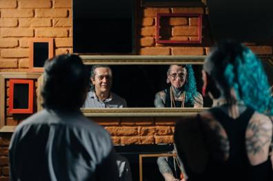 Foto de uma cena do documentário de Sérgio. Sérgio está ao lado de T’angel. Sérgio é um homem indígena, de cabelos grisalhos e veste uma camisa social azul clara. T’angel é uma pessoa branca, de cabelos ondulados azuis e veste camiseta preta. Possui tatuagens visíveis nas costas, rosto e pescoço. Ambas as pessoas estão posicionadas de costas para a câmera e é possível observar seus rostos e bustos através da reflexão de um espelho. O espelho está pendurado em uma parede de tijolos laranjas, que também possui espelhos de outras formas e tamanhos.