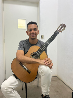 Fotografia de um homem jovem e pardo de cabelos curtos e escuros, sentado em uma sala de paredes brancas, segurando um violão sobre seu colo. Ele usa calças brancas e camiseta cinza, olha para a câmera e sorri, enquanto apoia a base do violão sobre suas pernas e passa os braços em volta do instrumento. Atrás dele há uma porta branca com uma pequena janela de vidro.