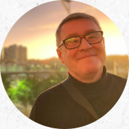 Foto de um homem branco sorrindo. Ele tem cabelos curtos e grisalhos, usa óculos de armação preta e blusa preta de gola alta. Ao fundo, o pôr do sol e prédios aparecem desfocados.
