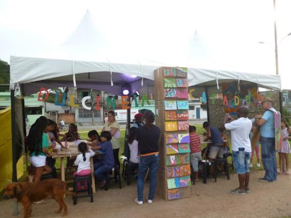 Foto de uma oficina cartonera. Há cerca de 15 crianças sentadas em bancos pretos junto a duas mesas ou de pé e quatro adultos observando a produção. As mesas estão debaixo de uma pequena tenda, com letreiros de papelão formando o texto “DULCINEIA CATADORA”. Na frente, há uma estante de livros cartoneros. O chão é de terra e ao fundo há uma rua residencial.