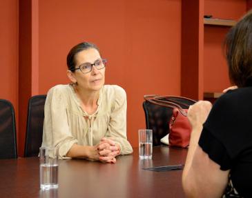 Foto da visita da professora Isabelle Millon Zumstein. Na imagem, a professora está ao centro, com as mãos cruzadas apoiadas numa mesa de madeira, prestando atenção na fala de uma mulher à sua frente. A professora é branca e tem cabelos castanhos presos em um coque baixo. Ela usa uma blusa bege e óculos de grau com armação azul. À sua frente, de costas, há uma mulher falando. Ela tem cabelo curto castanho e usa uma blusa preta. Ao lado da professora Isabelle ainda há uma bolsa vermelha e na mesa estão dois copos de vidro transparente com água.