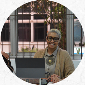 Foto de uma mulher negra de cabelos curtos e loiros, sentada e sorrindo para a câmera. Ela usa uma camisa cinza e sobretudo bege, óculos e um colar redondo texturizado em cores verde e dourado. Na mesa à sua frente, dois livros estão abaixo de um computador com alguns fios conectados na lateral da tela. Sua mãos estão posicionadas ao lado do computador e em uma delas ela segura uma caneta. Do lado do computador, parte de uma bolsa aparece na imagem. Atrás da mulher, janelas transparentes com grandes mostram o lado de fora da sala.
