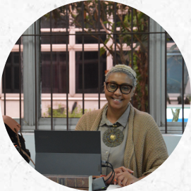 Foto de uma mulher negra de cabelos curtos e loiros, sentada e sorrindo para a câmera. Ela usa uma camisa cinza e sobretudo bege, óculos e um colar redondo texturizado em cores verde e dourado. Na mesa à sua frente, dois livros estão abaixo de um computador com alguns fios conectados na lateral da tela. Sua mãos estão posicionadas ao lado do computador e em uma delas ela segura uma caneta. Do lado do computador, parte de uma bolsa aparece na imagem. Atrás da mulher, janelas transparentes com grandes mostram o lado de fora da sala. 