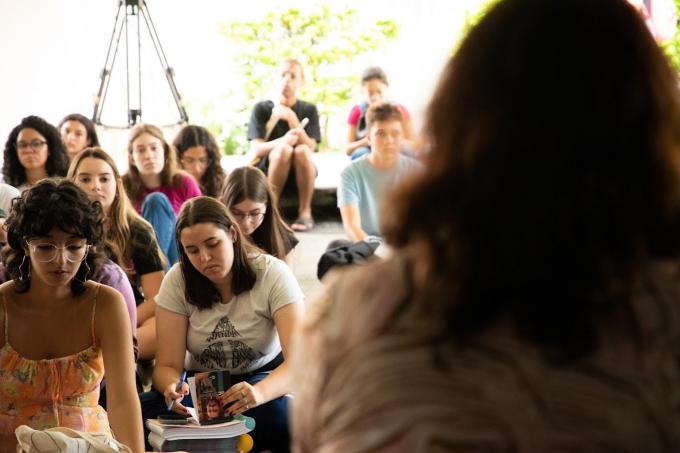 Fotografia de estudantes em uma palestra. Em primeiro plano, em desfoque no lado direito da imagem, há as costas de uma mulher. Ela usa camiseta estampada e tem cabelos castanhos e na altura dos ombros. Em segundo plano, no lado esquerdo da imagem, há cerca de 10 estudantes sentados de frente para a palestrante, sendo a maioria mulheres brancas. Ao fundo, há um espaço aberto, claro e com plantas.