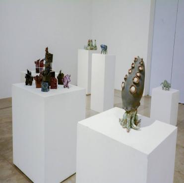 Foto de salão com pedestais onde estão expostas esculturas em cerâmica. No primeiro plano, há uma escultura de lula, e no fundo há um conjunto de pequenas esculturas distribuídas em outros três pedestais, e à esquerda, há um pedestal menor com apenas uma escultura de tartaruga.