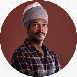 Foto de um homem negro, olhos escuros e bigode escuro que posa para a camêra. Ele usa uma camisa xadrez com fundo azul escuro e linhas alaranjadas. Ele usa um turbante de tecido cinza com uma parte branca no topo. O fundo da imagem é marrom.
